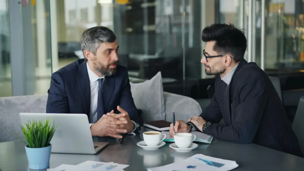 Two businessmen having a meeting with laptops, papers, and coffee at a modern office.