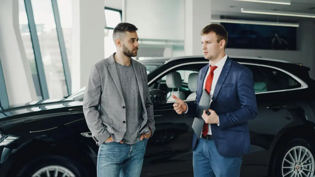 Two men discussing business in a car dealership, standing near a vehicle.